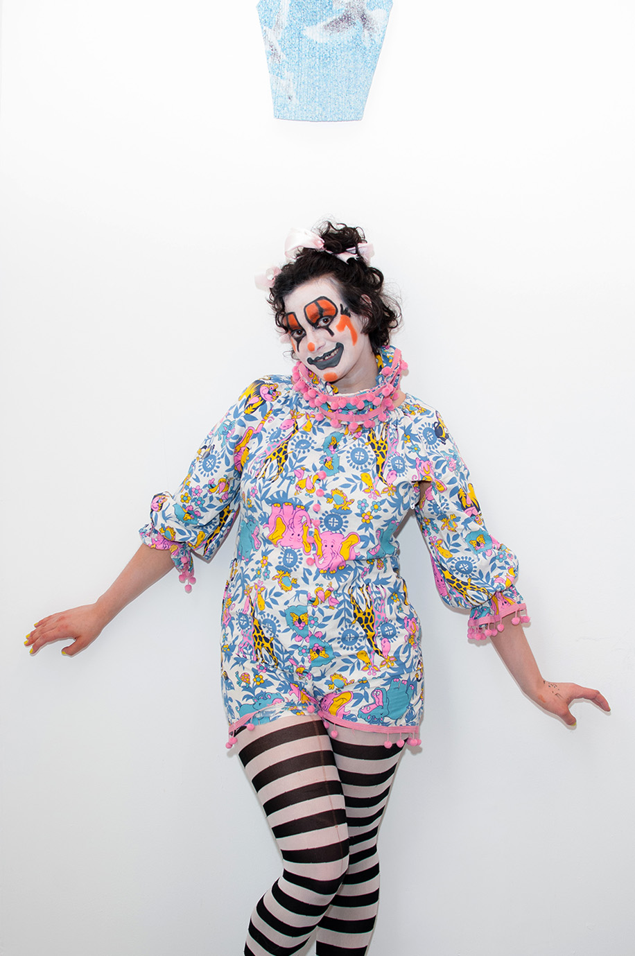 Person dressed as a clown poses under a pleated tank top hung on the wall